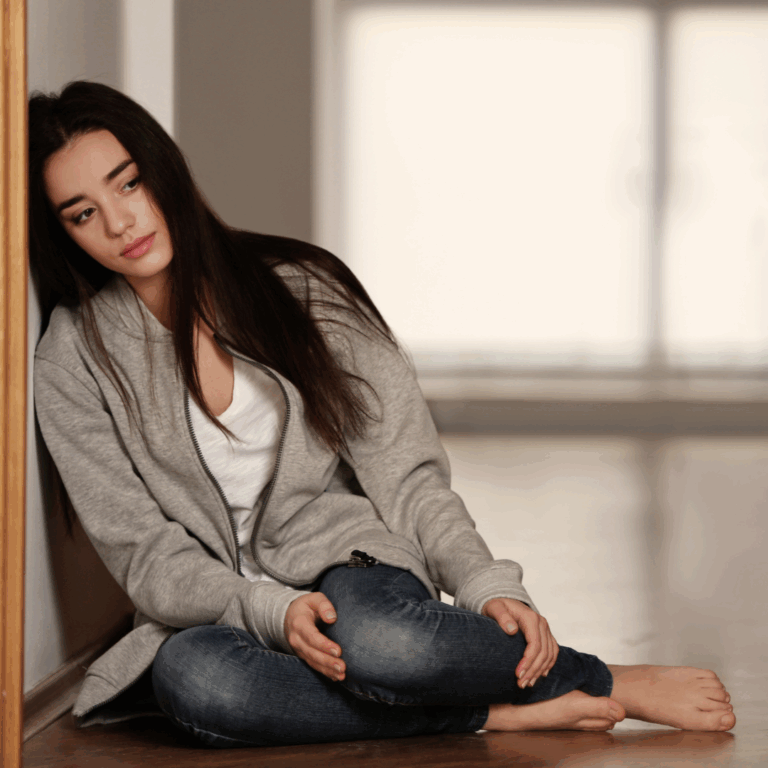 woman sitting on the floor, leaning against the wall looking off to the side with a sad expression on her face
