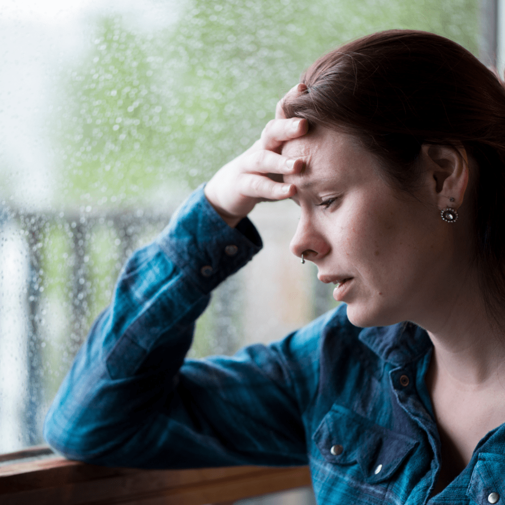 woman with elbow on windowsill, hand to forehead, looking overwhelmed
