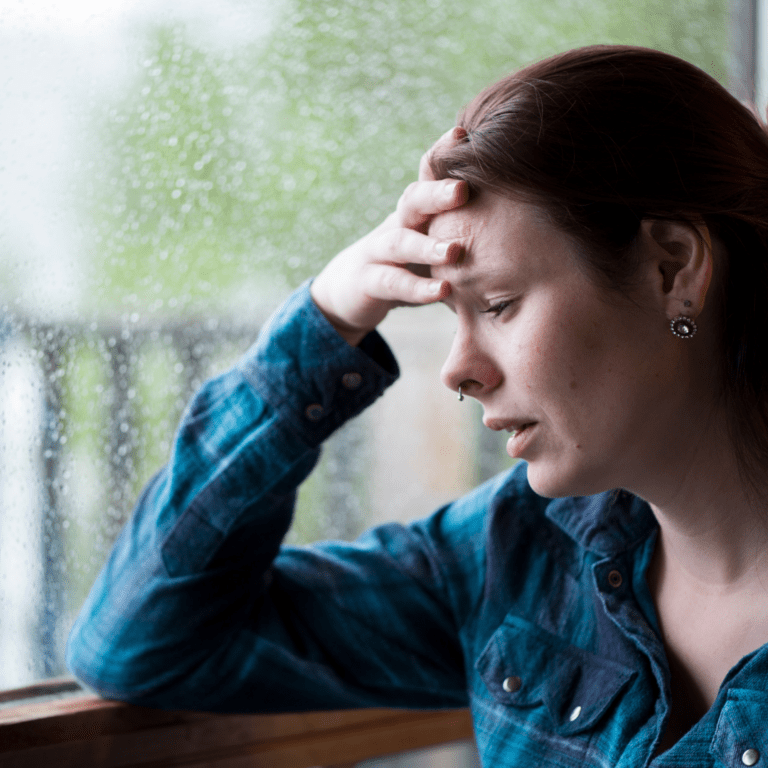 woman with elbow on windowsill, hand to forehead, looking overwhelmed